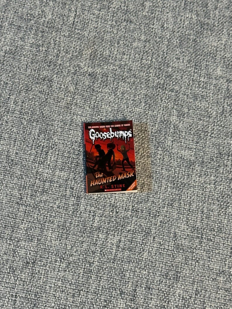 May include: A paperback book titled "Goosebumps: The Haunted Mask" by R.L. Stine. The cover features a dark red background with silhouetted figures and the title in a spooky font. The book is set against a gray textured surface.