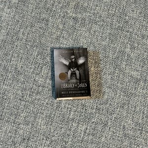 May include: A hardcover book titled "Library of Souls" by Ransom Riggs. The cover features a black and white image of a person with wings. The book is part of the "Miss Peregrine's Peculiar Children" series.