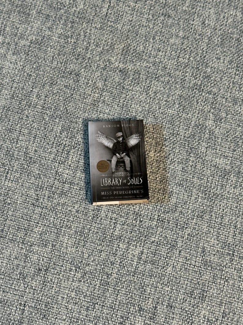 May include: A hardcover book titled "Library of Souls" by Ransom Riggs. The cover features a black and white image of a person with wings. The book is part of the "Miss Peregrine's Peculiar Children" series.