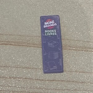 May include: A small, rectangular purple toy package with the words "Mini Brands" and "Books Livres" printed in white. The package has illustrations of books and a glossy finish.