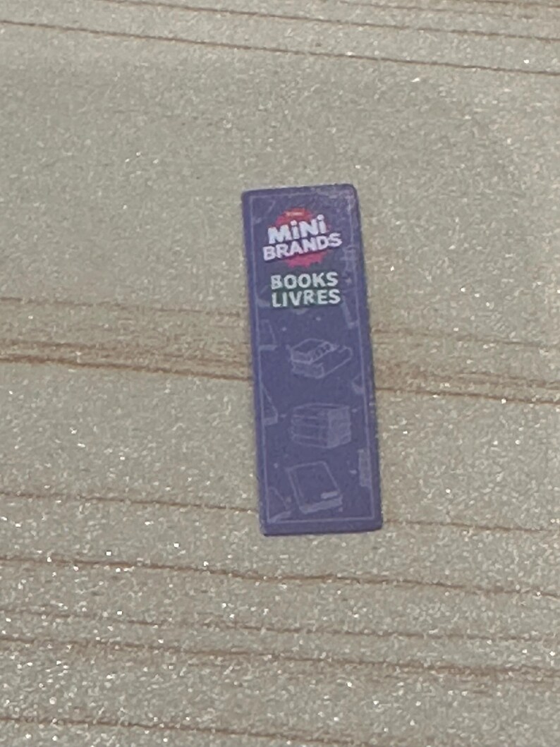 May include: A small, rectangular purple toy package with the words "Mini Brands" and "Books Livres" printed in white. The package has illustrations of books and a glossy finish.