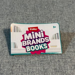 May include: A rectangular box for ZURU Mini Brands Books. The box has a white background with a pink and red starburst design that says "Mini Brands Books". The box has a teal border and the number "3+" in the upper right corner.