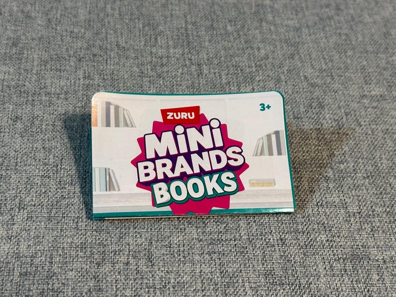 May include: A rectangular box for ZURU Mini Brands Books. The box has a white background with a pink and red starburst design that says "Mini Brands Books". The box has a teal border and the number "3+" in the upper right corner.