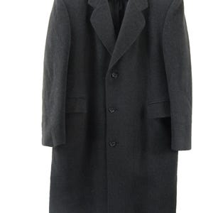 Può includere: Un cappotto lungo grigio scuro, al ginocchio, con revers a lancia e chiusura a tre bottoni. Il cappotto ha due tasche anteriori ed è appeso a una gruccia bianca. Il cappotto è realizzato in materiale simile alla lana.