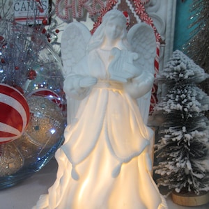 May include: A white ceramic angel figurine holding a harp, with outstretched wings. The angel's dress is illuminated from within, creating a soft glow. The base of the figurine has a cloud-like design. A Christmas decoration.