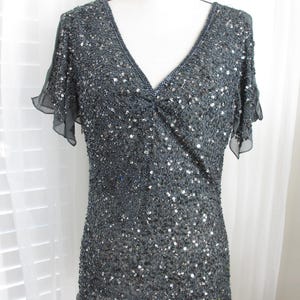 Puede incluir: Una blusa gris oscuro con lentejuelas, cuello en V y mangas cortas y transparentes. La blusa est&aacute; cubierta de peque&ntilde;as lentejuelas reflectantes, creando un efecto brillante. La prenda se exhibe en un maniqu&iacute;, mostrando su forma y dise&ntilde;o.