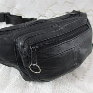 Op de afbeelding: Zwarte leren heuptas met meerdere vakken met ritssluiting. De tas heeft een zwarte verstelbare riem met een plastic gesp. De ritsen hebben metalen treklipjes. De tas is gemaakt van een getextureerd leer.