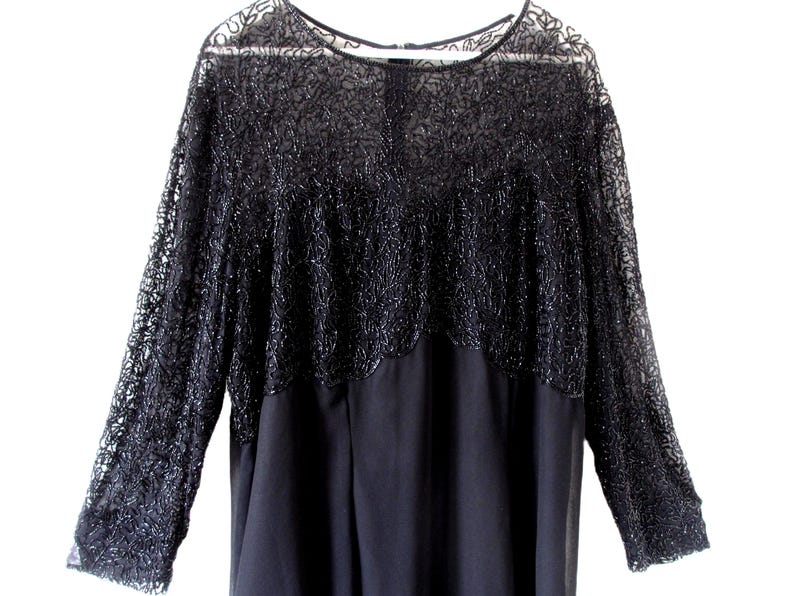 Puede incluir: Un vestido negro con mangas largas. La parte superior presenta un intrincado encaje con cuentas, mientras que la secci&oacute;n inferior es de tela oscura lisa. El vestido tiene un cuello redondo y una silueta fluida, adecuado para ocasiones formales.