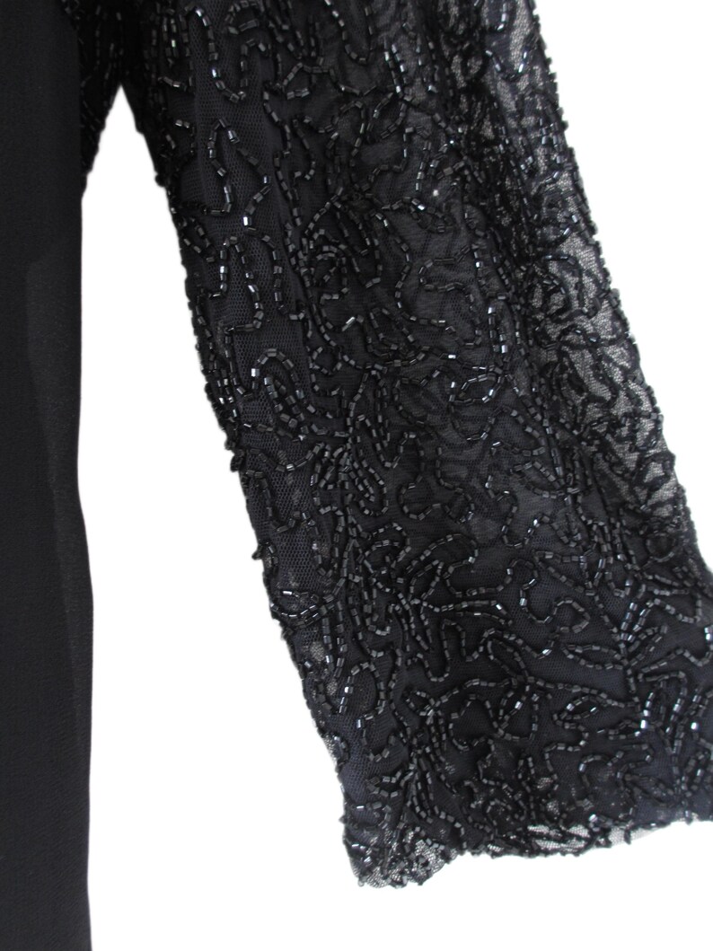 Puede incluir: Vestido negro con una manga adornada con abalorios. La manga es de tela transparente y est&aacute; cubierta con un intrincado patr&oacute;n de abalorios negros. El vestido en s&iacute; es de color negro liso, con la manga que proporciona un contraste decorativo.