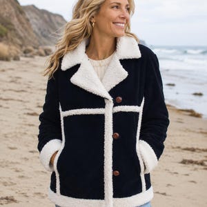 May include: Navy blue corduroy jacket with white faux shearling trim and lining. The jacket features a notched collar, three brown buttons, and two front pockets. The model is wearing blue jeans and is standing on a sandy beach.