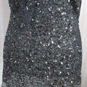 Puede incluir: Una prenda gris oscuro, probablemente una blusa o vestido, densamente cubierta de peque&ntilde;as lentejuelas y cuentas. La tela es un material transparente, con un dobladillo festoneado. El dise&ntilde;o crea un efecto brillante y texturizado.