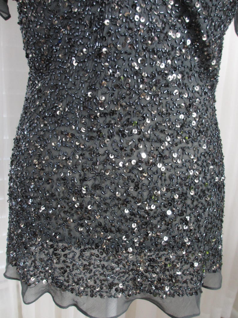 Puede incluir: Una prenda gris oscuro, probablemente una blusa o vestido, densamente cubierta de peque&ntilde;as lentejuelas y cuentas. La tela es un material transparente, con un dobladillo festoneado. El dise&ntilde;o crea un efecto brillante y texturizado.