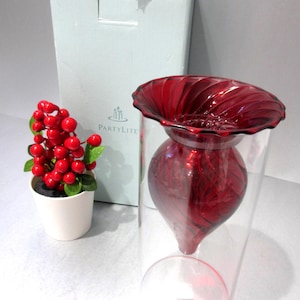 May include: A red glass candle holder with a decorative top and a clear glass base. A small white pot with red berries and green leaves is to the left. A white box is in the background.