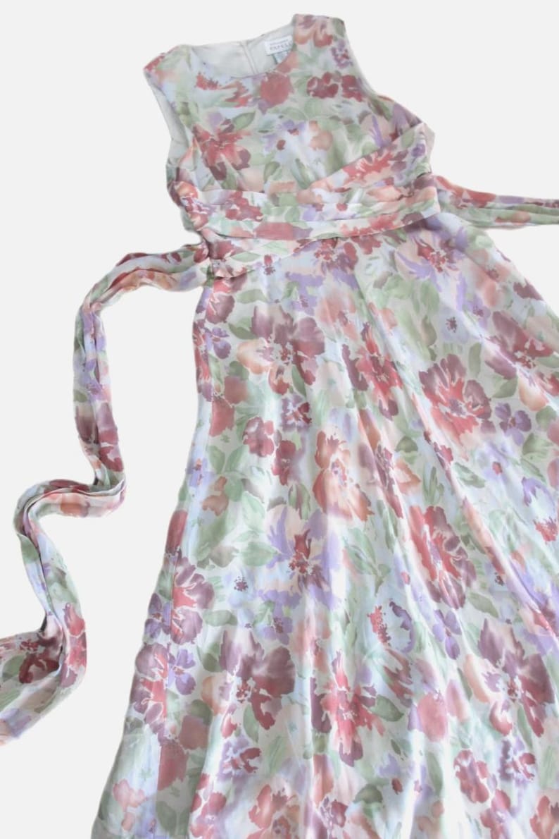 May include: A sleeveless floral print dress in shades of red, purple, and green on a white background. The dress has a flowing skirt and a matching sash that ties at the waist. The fabric appears to be lightweight.
