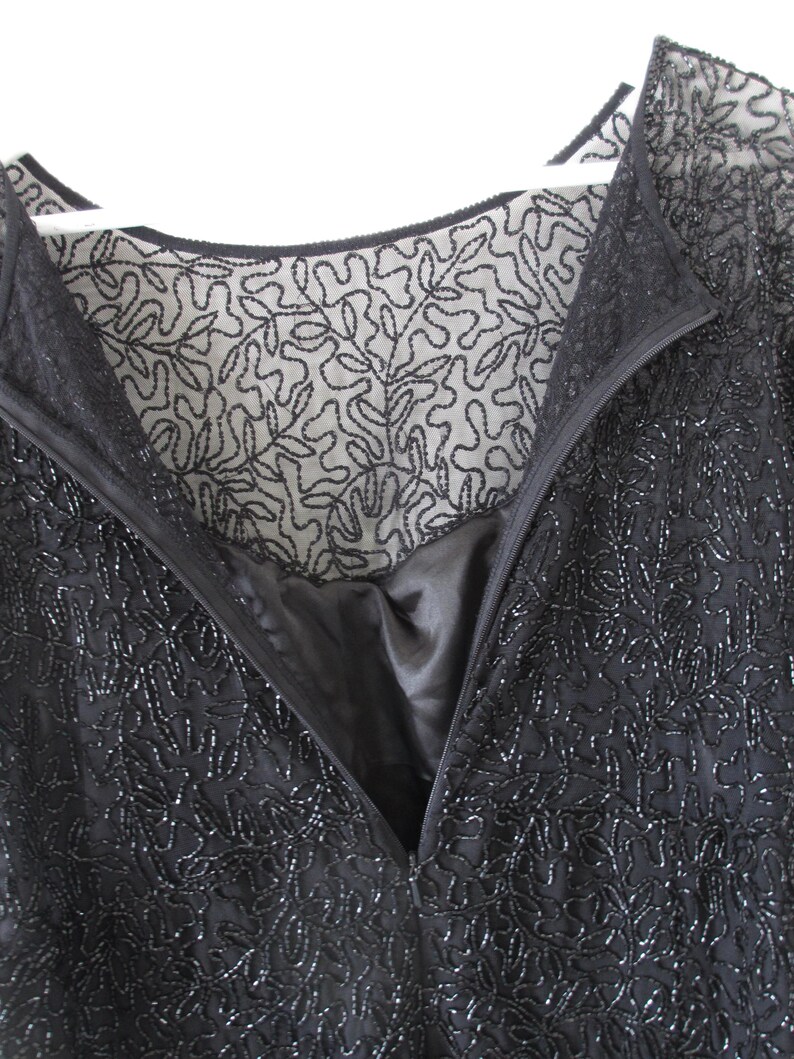 Puede incluir: Primer plano de un vestido negro con cuentas, con escote en V y cremallera. El vestido presenta intrincados patrones florales y un forro transparente bordado. El vestido est&aacute; colgado de una percha blanca.