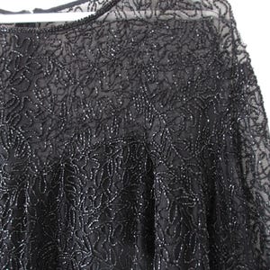 Puede incluir: Primer plano de un top negro con cuentas. El top presenta un intrincado trabajo de cuentas en un patr&oacute;n floral, con un escote y mangas transparentes. Las cuentas son de un color oscuro y reflectante, creando una apariencia texturizada.