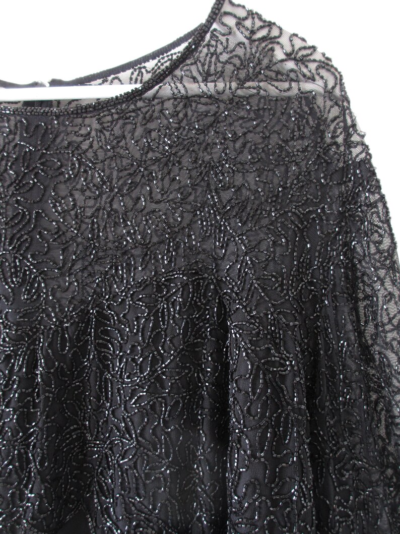 Puede incluir: Primer plano de un top negro con cuentas. El top presenta un intrincado trabajo de cuentas en un patr&oacute;n floral, con un escote y mangas transparentes. Las cuentas son de un color oscuro y reflectante, creando una apariencia texturizada.