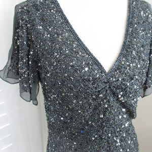 Puede incluir: Un vestido gris oscuro con escote en V y mangas cortas y transparentes. El vestido est&aacute; muy adornado con lentejuelas y cuentas, creando un efecto brillante. El escote y las mangas est&aacute;n ribeteados con cuentas m&aacute;s oscuras.