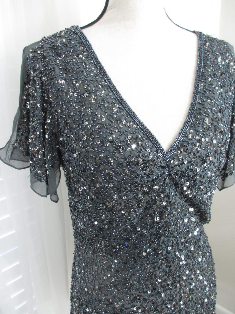 Puede incluir: Un vestido gris oscuro con escote en V y mangas cortas y transparentes. El vestido est&aacute; muy adornado con lentejuelas y cuentas, creando un efecto brillante. El escote y las mangas est&aacute;n ribeteados con cuentas m&aacute;s oscuras.