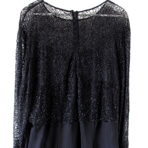 Puede incluir: Vestido de noche negro con mangas largas. La capa superior presenta un intrincado trabajo de cuentas sobre una tela transparente, creando un patr&oacute;n floral texturizado. El vestido tiene un cuello redondo y una cremallera en la espalda. La parte inferior es de tela negra lisa y opaca.