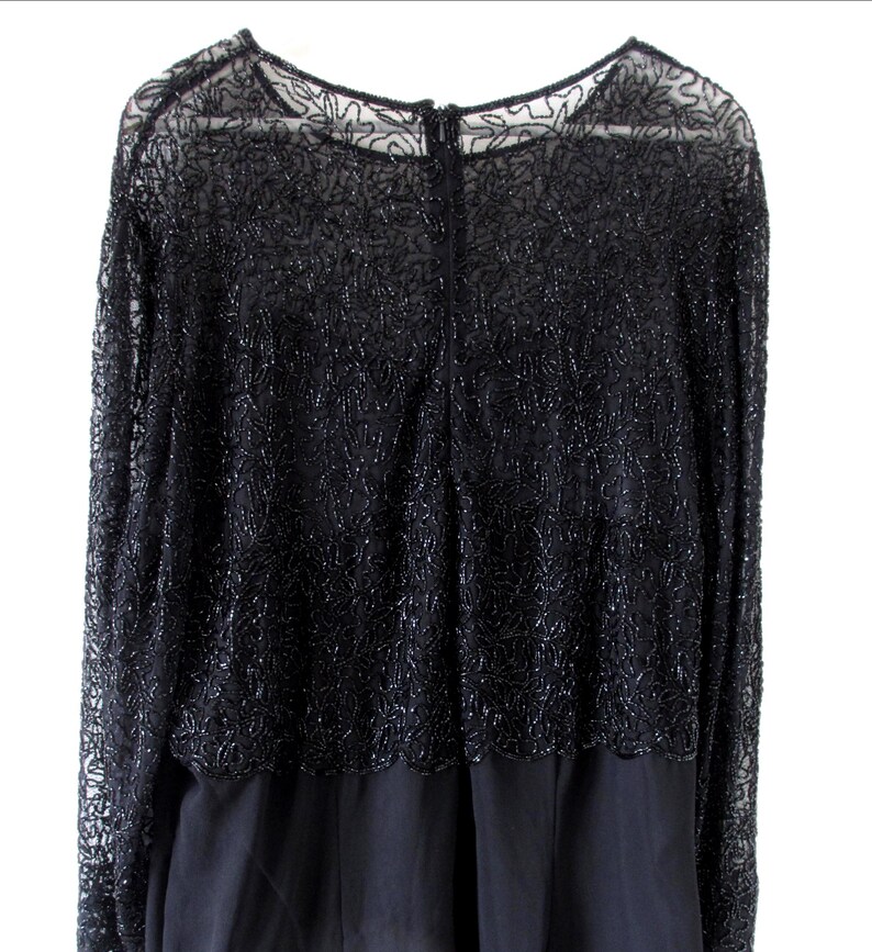 Puede incluir: Vestido de noche negro con mangas largas. La capa superior presenta un intrincado trabajo de cuentas sobre una tela transparente, creando un patr&oacute;n floral texturizado. El vestido tiene un cuello redondo y una cremallera en la espalda. La parte inferior es de tela negra lisa y opaca.