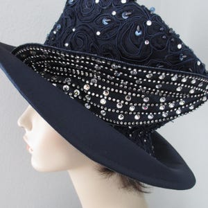 May include: A navy blue hat featuring a tall, structured crown adorned with intricate dark embroidery and scattered clear sequins. The wide, smooth brim is complemented by a decorative band densely encrusted with sparkling clear rhinestones.
