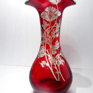 Puede incluir: Un jarrón de cristal rojo rubí con la parte superior acampanada y diseño floral plateado. El jarrón tiene una base redondeada y un borde plateado. El diseño floral plateado incluye flores y hojas. El jarrón mide unos 20 cm de alto.