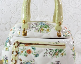May include: A white ceramic handbag-shaped container with a quilted pattern and floral designs in various colors. It features a light green handle and gold-colored accents. The container is sitting on a wooden surface.