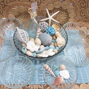 May include: A coastal-themed decorative arrangement featuring clear glass seashell-shaped bowls and a central bowl filled with seashells, stones, and decorative elements. A glass bottle and starfish add to the beachy aesthetic.