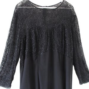 Puede incluir: Un vestido negro con mangas largas. La parte superior del vestido presenta una superposici&oacute;n de encaje con cuentas detalladas que se extiende hasta las mangas largas. La parte inferior del vestido es de tela lisa y uniforme. El vestido tiene un escote redondo.