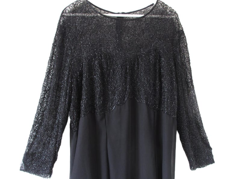 Puede incluir: Un vestido negro con mangas largas. La parte superior del vestido presenta una superposici&oacute;n de encaje con cuentas detalladas que se extiende hasta las mangas largas. La parte inferior del vestido es de tela lisa y uniforme. El vestido tiene un escote redondo.