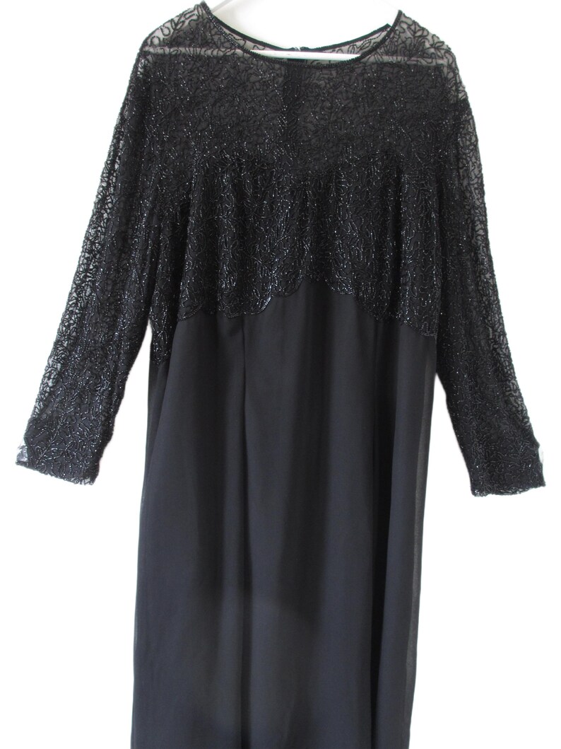 Puede incluir: Un vestido negro con mangas largas. La parte superior del vestido presenta un dise&ntilde;o detallado y texturizado con cuentas, mientras que la parte inferior es de tela lisa y fluida. El vestido cuelga de una percha blanca.