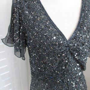 Puede incluir: Un vestido gris oscuro con cuentas, con escote en V y mangas cortas y transparentes. El vestido est&aacute; cubierto de peque&ntilde;as cuentas brillantes, creando una superficie texturizada. El escote y las mangas est&aacute;n ribeteados con un borde m&aacute;s oscuro y densamente adornado con cuentas.
