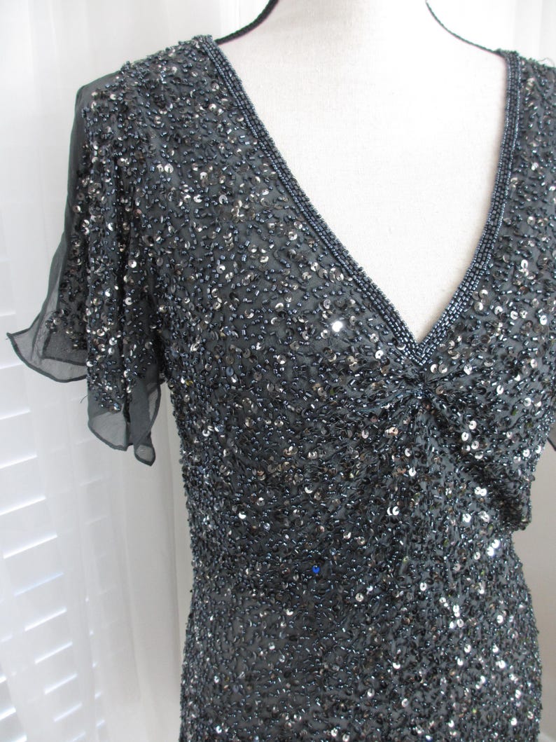 Puede incluir: Un vestido gris oscuro con cuentas, con escote en V y mangas cortas y transparentes. El vestido est&aacute; cubierto de peque&ntilde;as cuentas brillantes, creando una superficie texturizada. El escote y las mangas est&aacute;n ribeteados con un borde m&aacute;s oscuro y densamente adornado con cuentas.