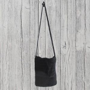 May include: A black crochet crossbody bag with a long strap, hanging against a light wood-grain background. The bag has a rectangular shape with a slightly wavy top edge and a darker patch on the front.