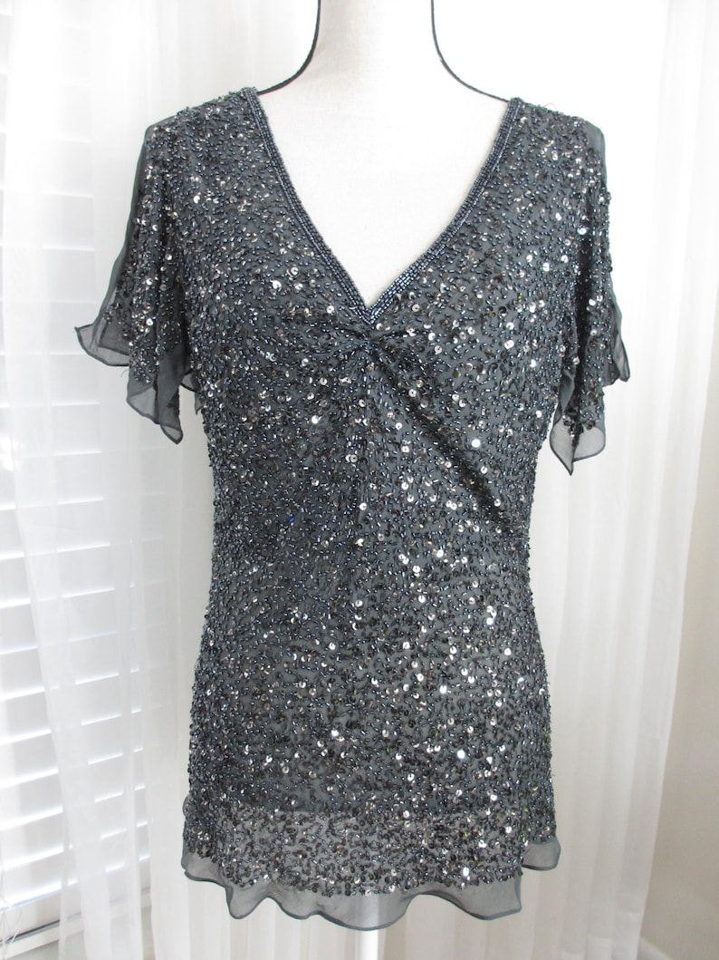 Puede incluir: Una blusa gris oscuro con lentejuelas, cuello en V y mangas cortas y transparentes. La blusa est&aacute; cubierta de peque&ntilde;as lentejuelas reflectantes, creando un efecto brillante. La prenda se exhibe en un maniqu&iacute;, mostrando su forma y dise&ntilde;o.