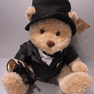 Peut inclure: Un ours en peluche portant un chapeau haut de forme noir et un smoking. L'ours a un nez brun et tient une petite cloche dorée avec un ruban noir sur lequel on peut lire "2000". Une étiquette sur l'ours indique "Millennium Wellington Bear".