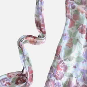 May include: A floral print dress with a flowing design. The dress features a pattern of purple, pink, and green flowers on a white background. The dress has a long, flowing scarf attached to the neckline.