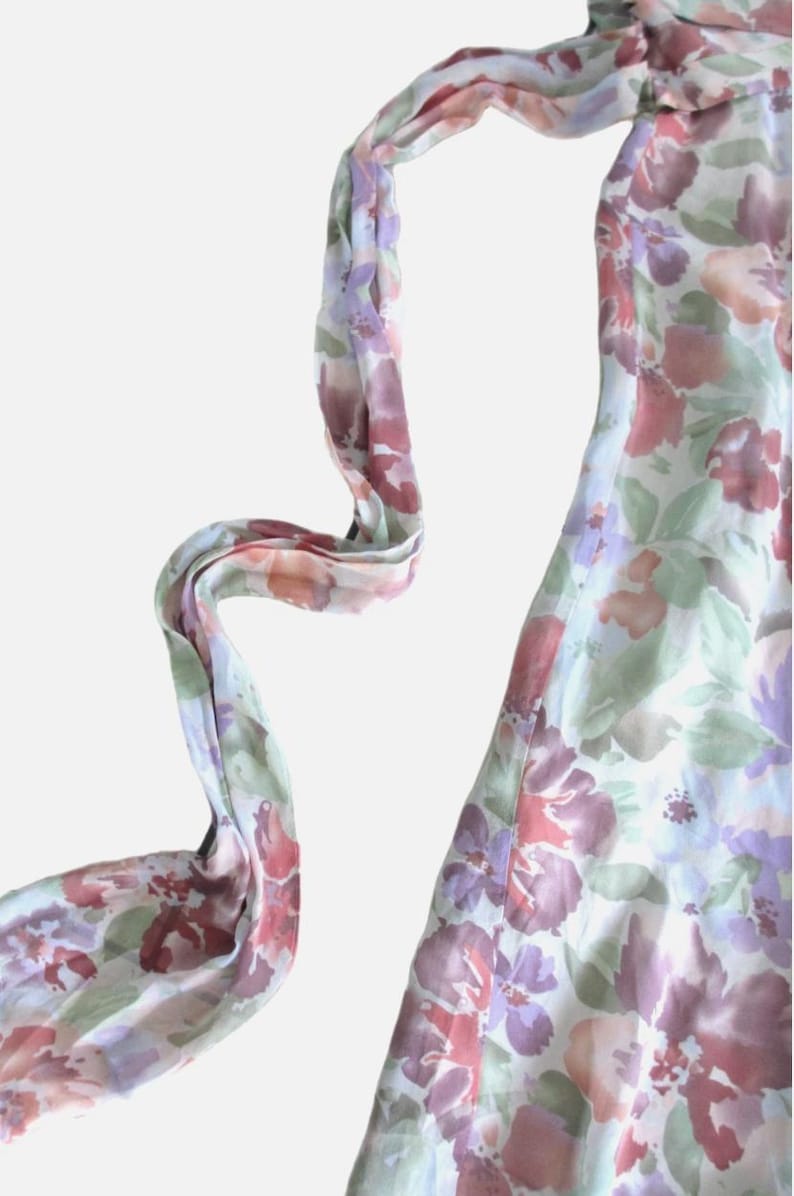 May include: A floral print dress with a flowing design. The dress features a pattern of purple, pink, and green flowers on a white background. The dress has a long, flowing scarf attached to the neckline.