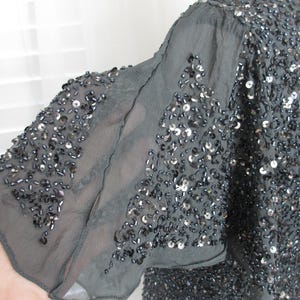 Puede incluir: Top gris oscuro con lentejuelas y mangas sueltas de gasa. La prenda est&aacute; ricamente adornada con peque&ntilde;as lentejuelas y cuentas reflectantes, creando un efecto brillante. Las mangas son de tejido delicado y semitransparente.