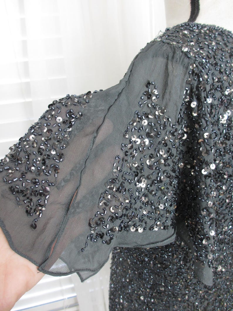Puede incluir: Top gris oscuro con lentejuelas y mangas sueltas de gasa. La prenda est&aacute; ricamente adornada con peque&ntilde;as lentejuelas y cuentas reflectantes, creando un efecto brillante. Las mangas son de tejido delicado y semitransparente.