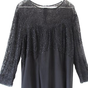 Puede incluir: Un vestido negro con mangas largas. La parte superior del vestido presenta intrincados detalles de cuentas y mangas de encaje transparente. La parte inferior del vestido es de tela negra lisa. El vestido est&aacute; colgado de una percha blanca.