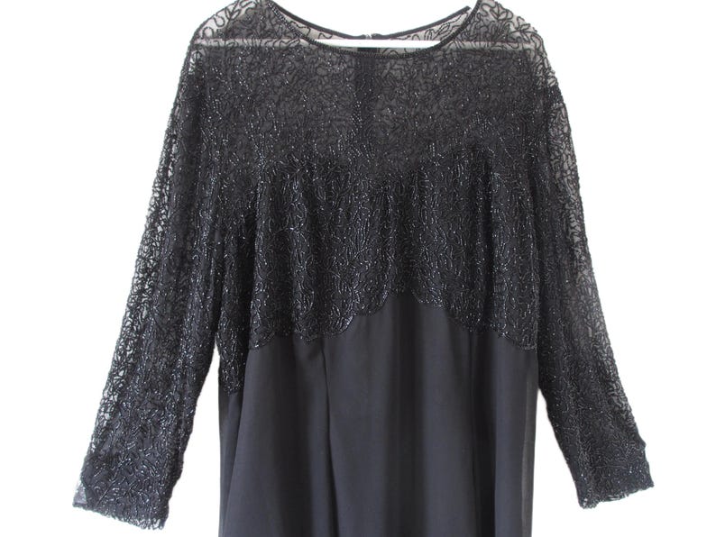Puede incluir: Un vestido negro con mangas largas. La parte superior del vestido presenta intrincados detalles de cuentas y mangas de encaje transparente. La parte inferior del vestido es de tela negra lisa. El vestido est&aacute; colgado de una percha blanca.