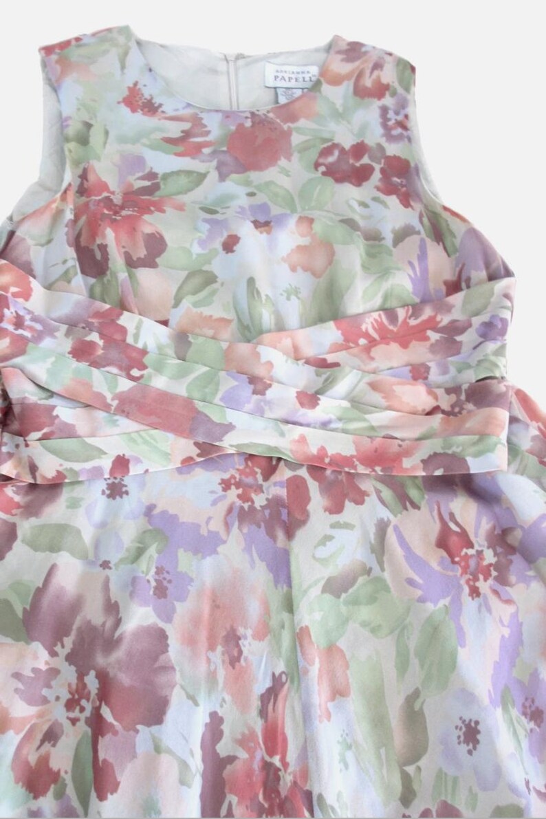 May include: A sleeveless floral dress featuring a watercolor-style print in shades of pink, purple, and green. The dress has a cinched waist detail and a flowing skirt. The fabric appears lightweight and suitable for warmer weather.