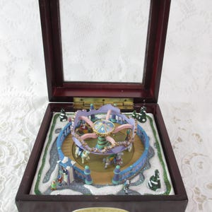 May include: A dark wood music box with a hinged glass lid containing a miniature carousel scene. The carousel features pink and blue structural elements, small horses, and figures on a snowy base. A gold plaque on the front reads Merry Go Round.