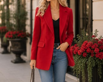 May include: A woman wearing a bright red blazer over a black top and blue jeans, holding a black quilted handbag. The blazer has a classic tailored design. The background features a building with decorative elements and red flowers.