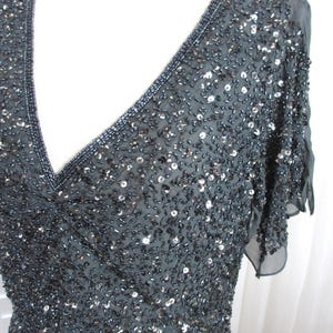 Puede incluir: Un vestido gris oscuro con lentejuelas, escote en V y mangas cortas y transparentes. El vestido est&aacute; muy adornado con lentejuelas y abalorios, creando un efecto brillante. El escote y las mangas est&aacute;n ribeteados con abalorios m&aacute;s oscuros.