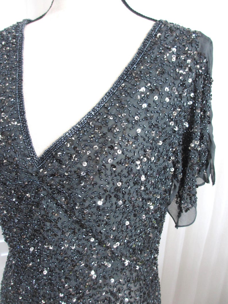 Puede incluir: Un vestido gris oscuro con lentejuelas, escote en V y mangas cortas y transparentes. El vestido est&aacute; muy adornado con lentejuelas y abalorios, creando un efecto brillante. El escote y las mangas est&aacute;n ribeteados con abalorios m&aacute;s oscuros.