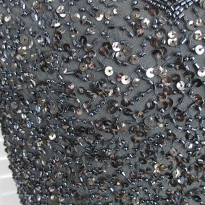 Puede incluir: Primer plano de un top con lentejuelas gris oscuro. La tela est&aacute; densamente cubierta de peque&ntilde;as lentejuelas redondas de color gris plateado y peque&ntilde;as cuentas. El escote est&aacute; adornado con hileras de peque&ntilde;as cuentas oscuras. El efecto general es una superficie brillante y texturizada.