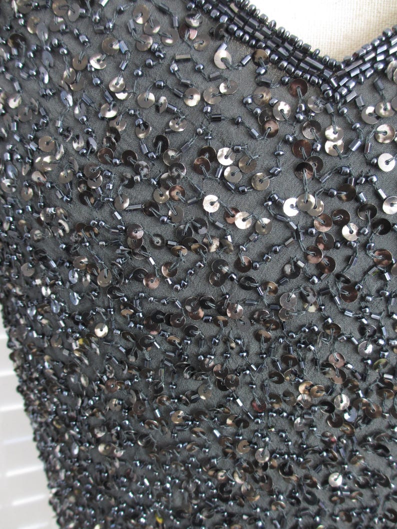 Puede incluir: Primer plano de un top con lentejuelas gris oscuro. La tela est&aacute; densamente cubierta de peque&ntilde;as lentejuelas redondas de color gris plateado y peque&ntilde;as cuentas. El escote est&aacute; adornado con hileras de peque&ntilde;as cuentas oscuras. El efecto general es una superficie brillante y texturizada.
